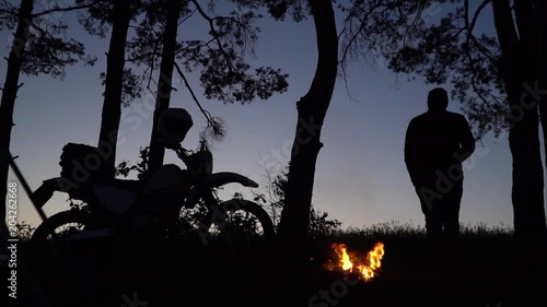 The silhouette of a cross off road motorcycle on a background of forest part in the evening, tent camp fire, bonfire, the man throws wood on the fire, and drinks tea, coffee from a cup, slow motion