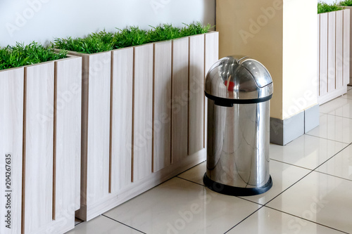 stainless steel trash can stands near the fence with greenery