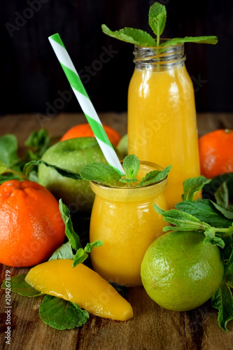 Yellow smoothie in glass jars surrounded by fruits and mint against wooden background