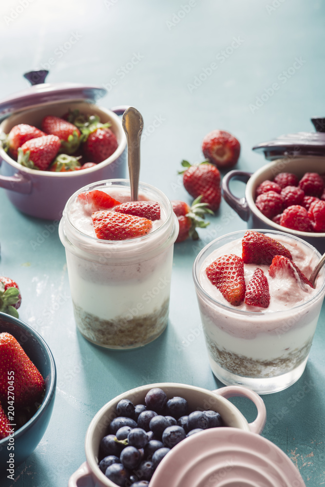 Strawberry cheesecake inside glass jar over a blue background and top view of berries , inside ceramic colored cocotte, blueberries, strawberries, raspberries, flat lay Strawberry cheesecake inside glass jar over a blue background and top view of berries , inside ceramic colored cocotte, blueberries, strawberries, raspberries, flat lay