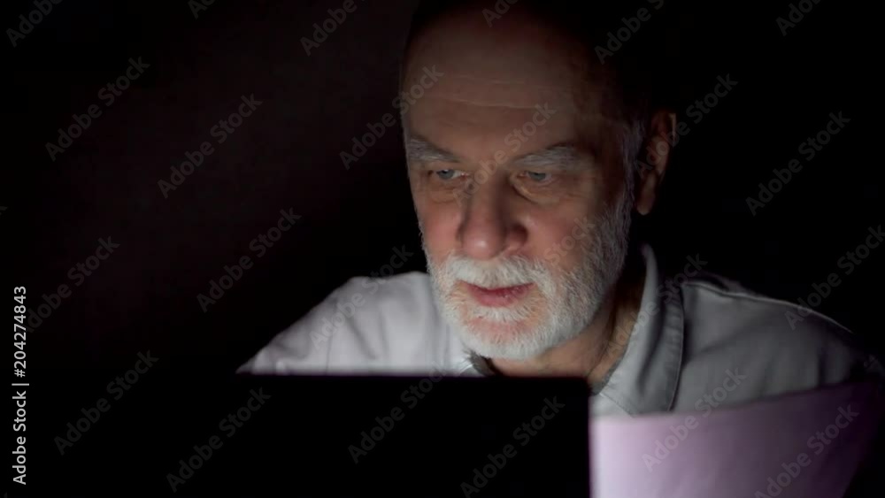 Tired overworked businessman at home working on laptop with documents late at night. Checking data for annual financial report. Secretly using computer hiding from people. Dark only face illuminated Tired overworked businessman at home working on laptop with documents late at night. Checking data for annual financial report. Secretly using computer hiding from people. Dark only face illuminated