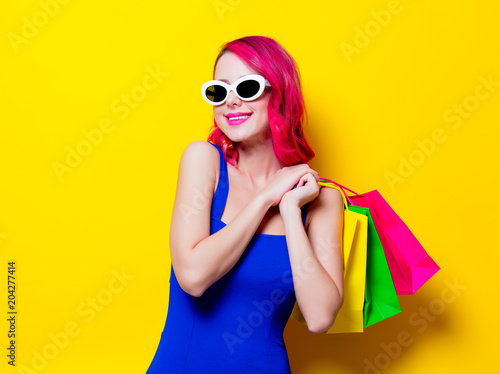 Young pink hair girl in blue dress with colored shopping bags. Portrait isolated on yellow background
