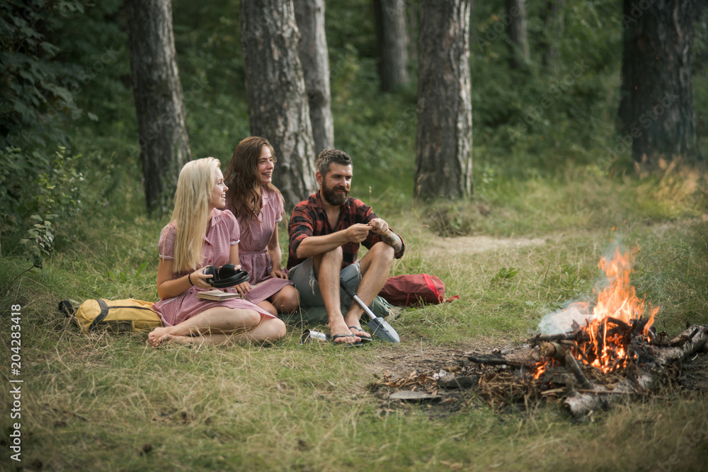 Happy friends enjoying picnic in woods. Bearded man and two sisters in retro dresses sitting next to campfire and cooking food outdoors Happy friends enjoying picnic in woods. Bearded man and two sisters in retro dresses sitting next to campfire and cooking food outdoors