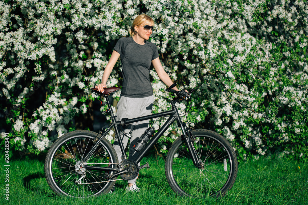 Pretty blonde woman in sunglasses standing with bicycle near blooming tree. Female cyclist in park