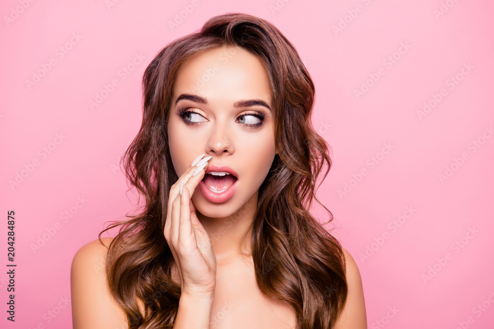 Fototapeta premium Close up portrait of cute charming gossip naked girl with wavy hair and trendy, modern make up holding hand near mouth and telling secret, looking at side with eyes, standing over pink background