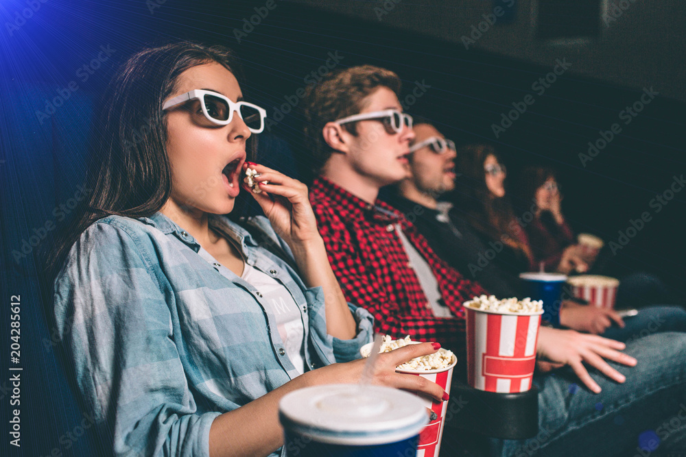 Fototapeta premium Amazed and surprised people are sitting in one row and watching movie. Blonde girl is eating popcorn with amazement on her face because of movie. The other people are amazed as well.