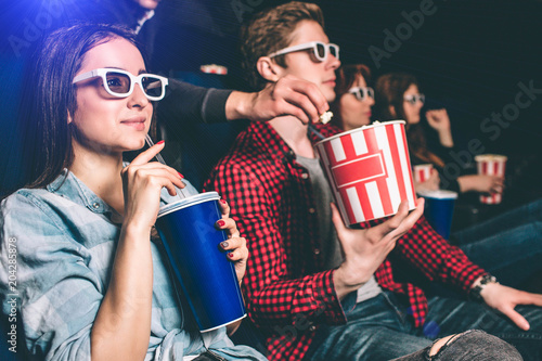 Somebody is reaching to the basket with popcorn that guy has and trying to take some popcorn from it. Guy doesn't see it because he is watching movie. Girl doesn't see it too. She holds a cup of coke.