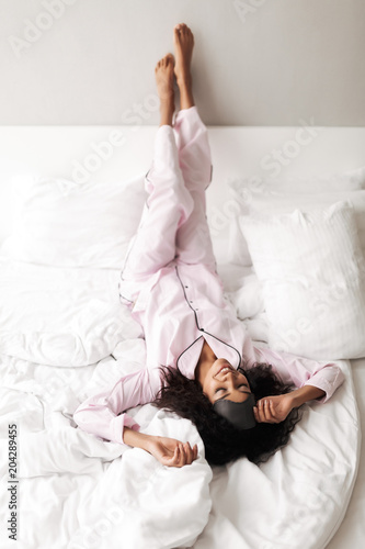 Portrait of young lady with dark curly hair lying in bed with eye mask on head and dreamily closing her eyes isolated