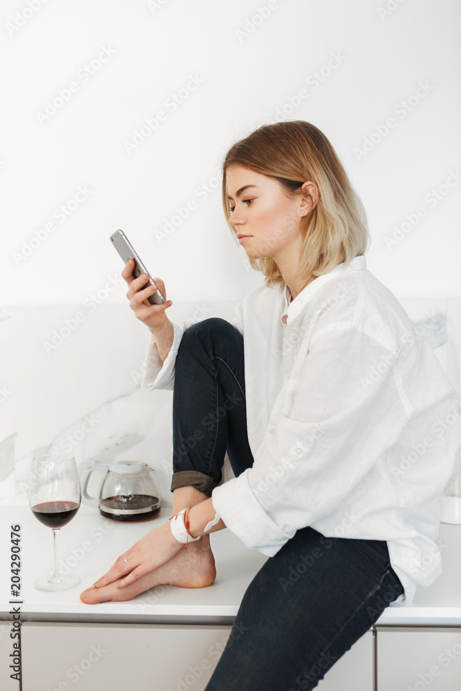 Portrait of young lady sitting on kitchen counter with cellphone in hand and glass of red wine near at home isolated Portrait of young lady sitting on kitchen counter with cellphone in hand and glass of red wine near at home isolated