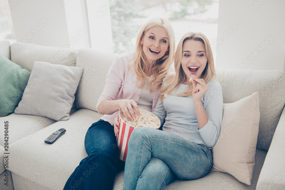 Cheerful laughing excited positive mother and daughter eating pop-corn looking tv, television sitting in living room indoor watching funny comedy, comfort domestic lifestyle free time Cheerful laughing excited positive mother and daughter eating pop-corn looking tv, television sitting in living room indoor watching funny comedy, comfort domestic lifestyle free time