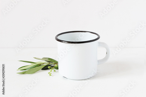 Styled stock photo. Feminine still life composition with blank white metal coffee enamel mug and green olive branch on white table background. Summer rustic scene, product mockup.