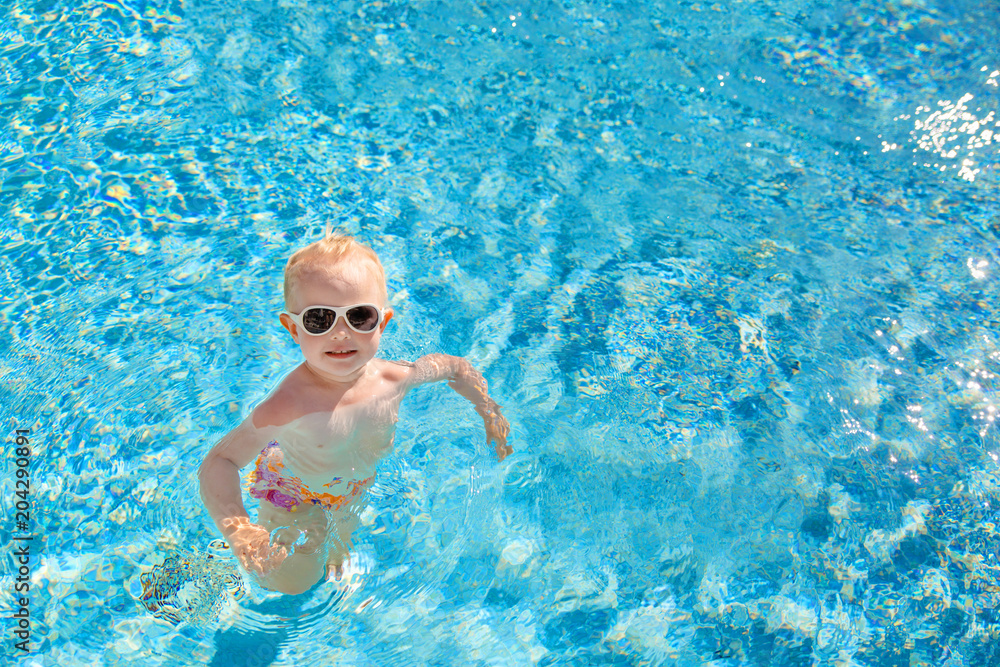 Little blonde girl swims in the pool with blue water. The view from the top. Place for text.