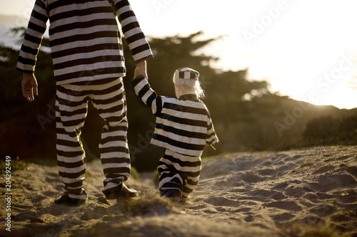 Young boy walking hand in hand with his father while wearing a striped prisoner uniform on a beach at sunset.