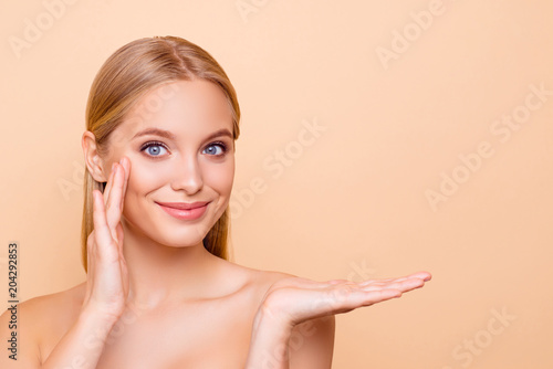 Pretty charming cute girl with perfect smooth soft skin holding copy space empty place on her palm applying product on her face with fingers, advertisement concept, isolated on beige background