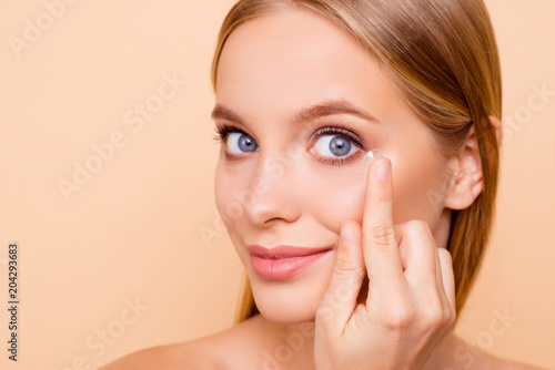 Fototapeta Naklejka Na Ścianę i Meble -  Portrait of pretty, charming, nude, natural cute, cheerful girl putting crystalline lens in left eye with hand, forefinger looking at camera isolated on beige background