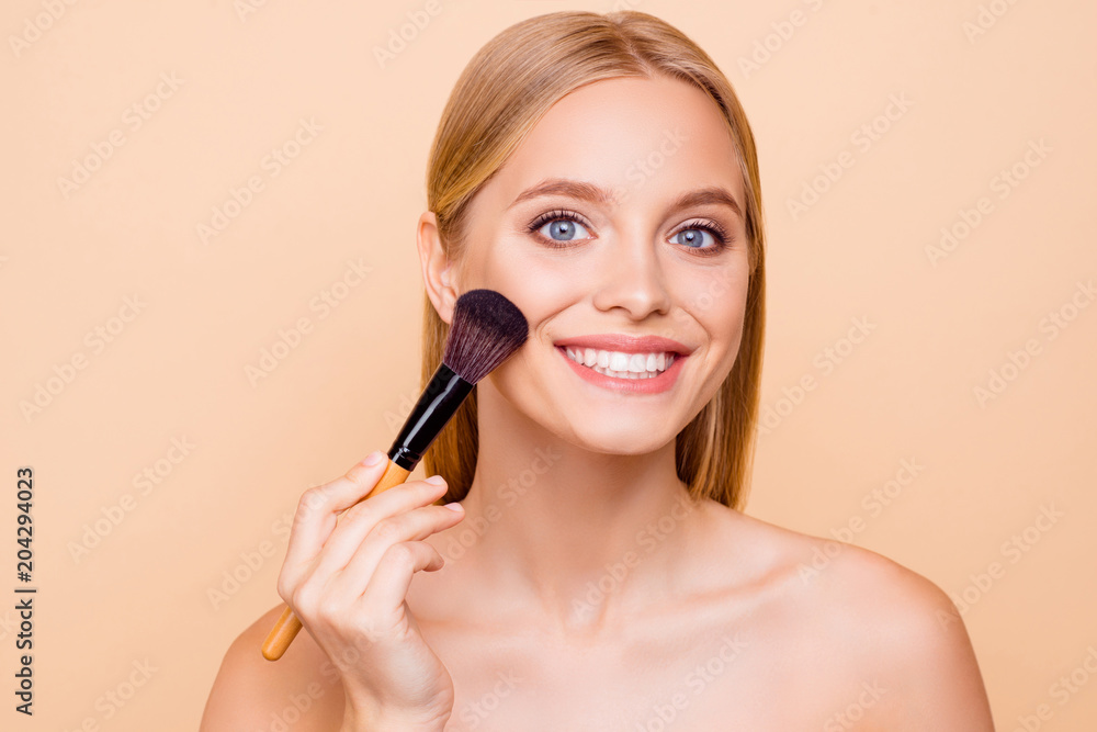 Charming pretty cheerful shine positive toothy woman applying foundation powder with makeup brush on cheek, having perfect smooth soft skin, looking at camera, isolated on beige background