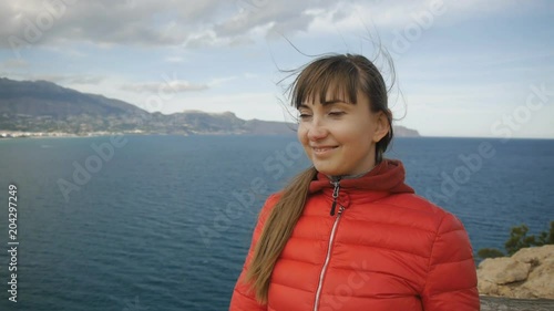 Young woman on sea background. Smiling caucasian brown-haired girl with fluttering hair in the wind looking forward from the mountain