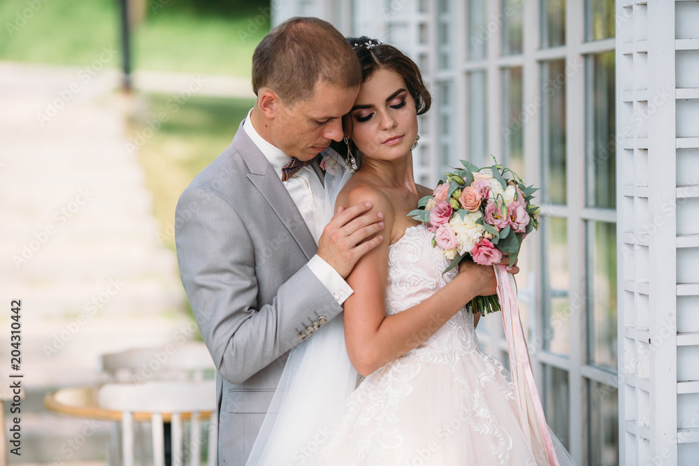 Portrait of newlyweds on wedding day. The groom in a gray suit with a white shirt and a bow tie hugs a beautiful bride with a luxurious white dress with a veil. Girl with makeup and hairdress with