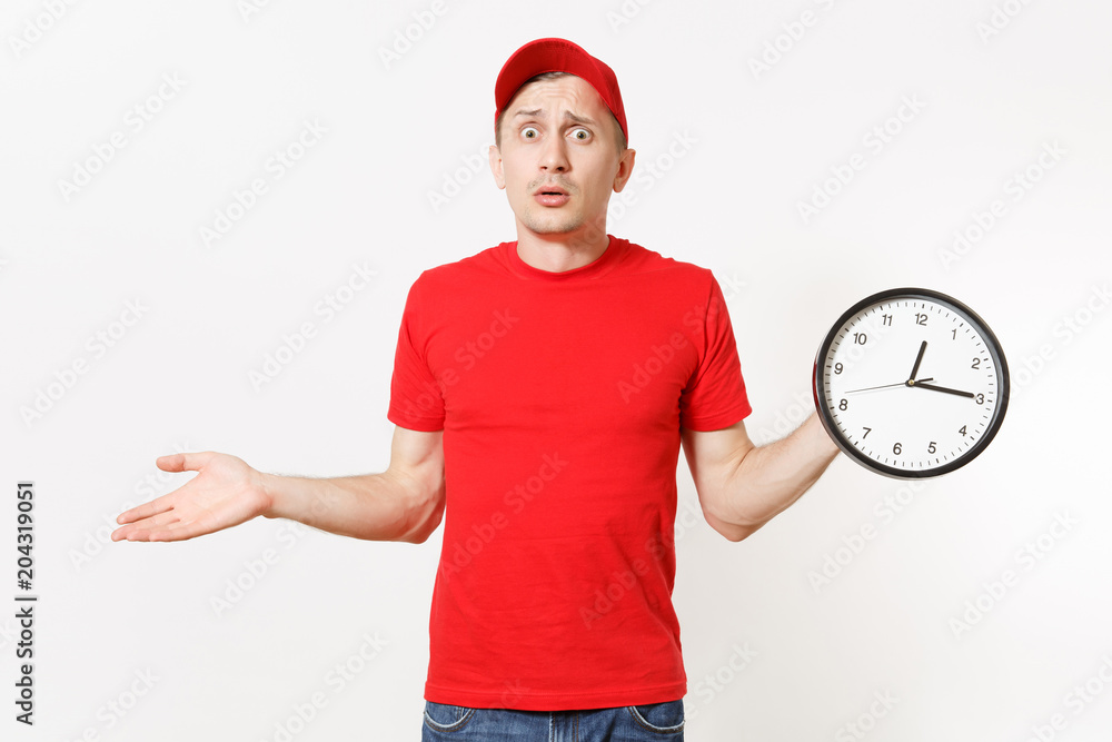 Delivery man in red uniform isolated on white background. Professional male in cap, t-shirt, jeans working as courier or dealer, holding round clock, showing in time. Copy space for advertisement. Delivery man in red uniform isolated on white background. Professional male in cap, t-shirt, jeans working as courier or dealer, holding round clock, showing in time. Copy space for advertisement.
