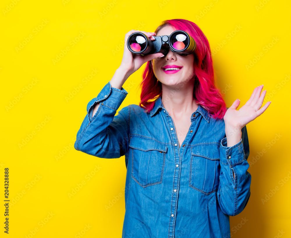 Young pink hair girl in blue shirt holding a binoculars. Portrait on isolated yellow background Young pink hair girl in blue shirt holding a binoculars. Portrait on isolated yellow background