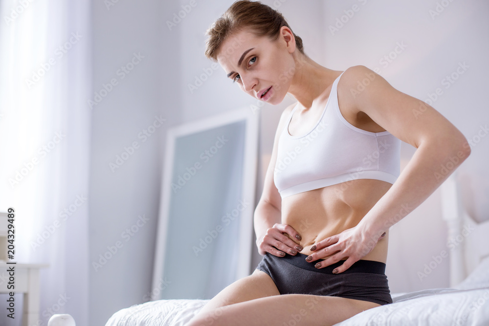 Weight control. Sad slim woman sitting on the bed while holding her waist Weight control. Sad slim woman sitting on the bed while holding her waist