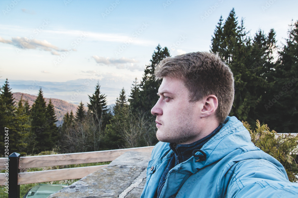 A young handsome blond guy thoughtfully looks into the distance against the background of mountains and coniferous trees, side view A young handsome blond guy thoughtfully looks into the distance against the background of mountains and coniferous trees, side view