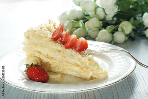 A piece of Napoleon cake on a plate adorned with a red ripe strawberry, a bouquet next to it