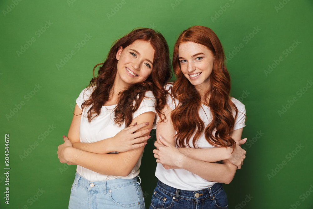 Portrait of two adorable girls 20s with ginger hair in casual wear smiling and looking at camera with arms folded, isolated over green background Portrait of two adorable girls 20s with ginger hair in casual wear smiling and looking at camera with arms folded, isolated over green background