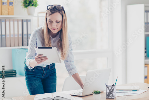 Portrait of charming, cute, busy woman having tablet in hand, having computer for work, using wi-fi internet, standing near desktop in work place, station photo