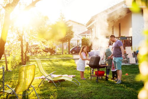 Beautiful couple and their adorable children have gathered around the grill  in front of the house.