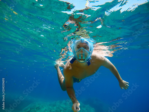 Underwater view of a young diver man swimming in the turquoise sea under the surface with snorkeling mask for summer vacation while taking a selfie with a stick.