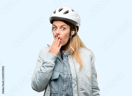 Young woman with bike helmet and earphones covers mouth in shock, looks shy, expressing silence and mistake concepts, scared