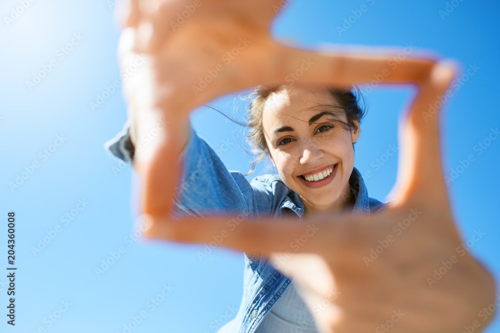 Naklejka premium portrait of a young smiling attractive woman in jeans clothes at sunny day on the blue sky background. woman shows a frame from hands like photo. Photo Frame Hands Made By A Hipster young girl