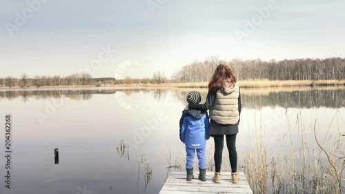 Mother and son are standing on small wooden pier at beautiful pond and enjoying countryside. Red-haired woman and child are enjoying nice lake. Sunny spring day, family values, happy atmosphere