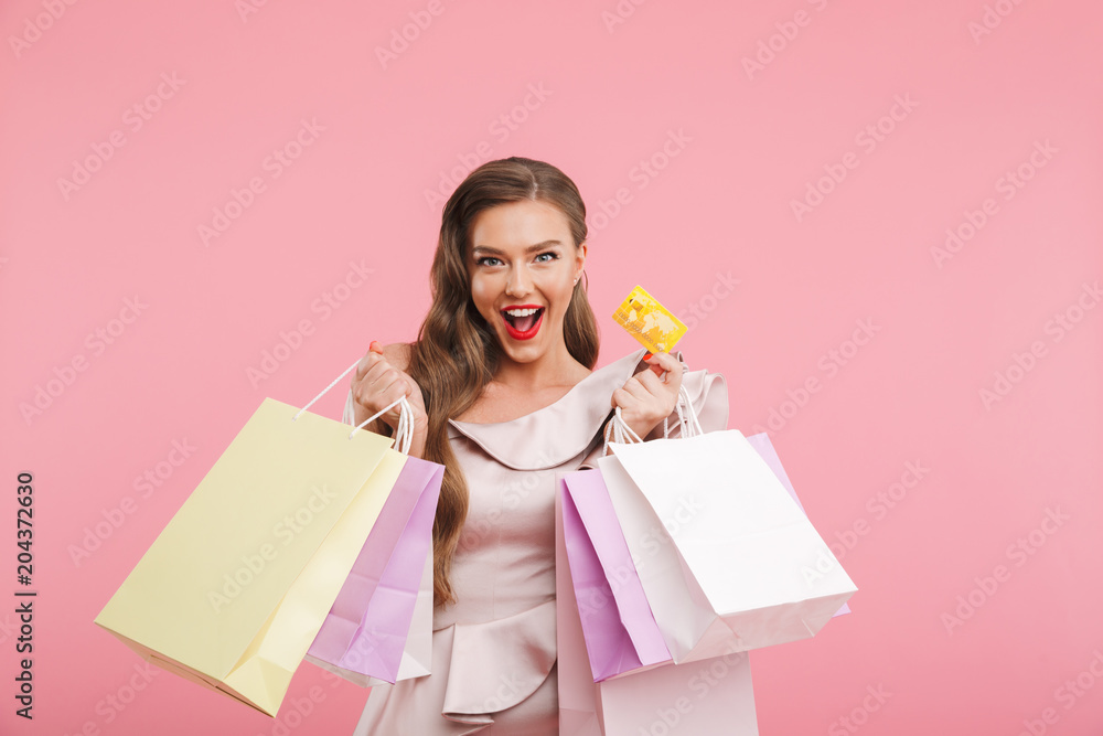 Fototapeta premium Photo of rich fashion woman 20s in dress smiling while holding different shopping bags and credit card in hands, isolated over pink background