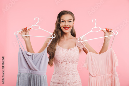 Photo of shopaholic woman 20s holding and choosing two dresses on hangers while shopping, isolated over pink background photo