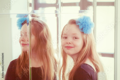 A girl with a blue bow in her hair stands near the mirror and looks at the camera and smiles