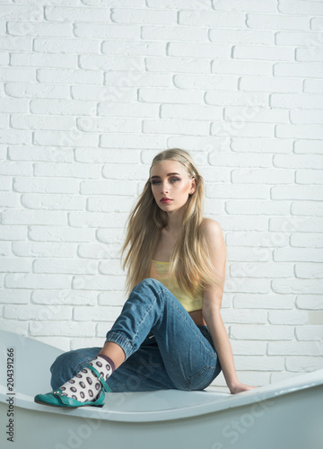 Blond model with messy hair sitting on the edge of bath tube in front of white brick wall