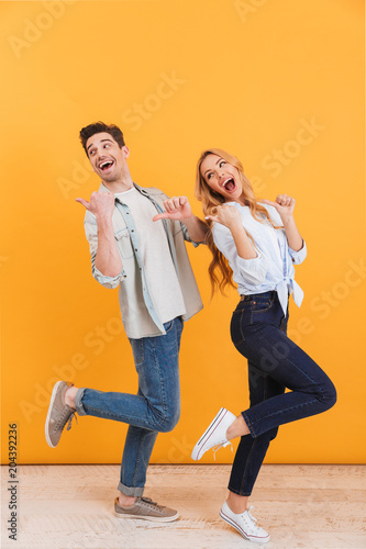 Full length photo of cheerful man and woman lookind back while pointing fingers at copyspace, isolated over yellow background