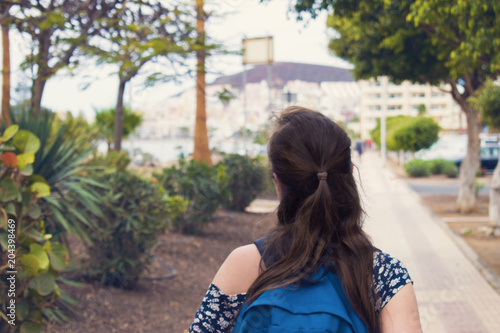 The young brown-haired woman in the printed t-shirt with the blue backpack walk around the resort  Playa de las Americas in Tenerife, Spain.  Back view.