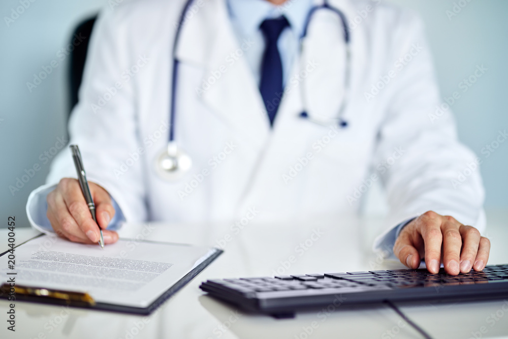 Male doctor works with computer and writes notes on the clipboard in the office Male doctor works with computer and writes notes on the clipboard in the office