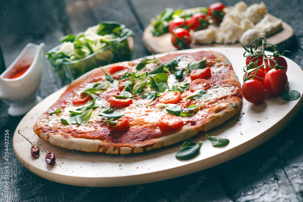 Italian food, cuisine. Margherita pizza on a black, wooden table with igredients like tomatoes, salad, cheese, mozzarella, basil. Delicious homemade food. 