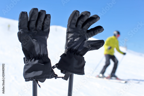 Detail on black ski gloves on ski poles with blurred skier in background on bright sunny winter day.