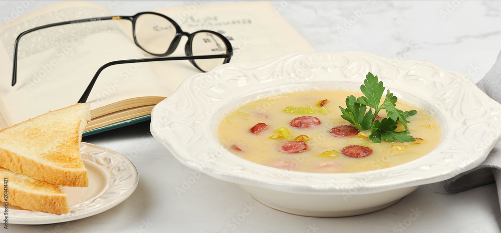 Potato soup with sausages in a deep dish. Next to the plate with toast of white bread. In the frame is a book and glasses. Close-up. Light background. Potato soup with sausages in a deep dish. Next to the plate with toast of white bread. In the frame is a book and glasses. Close-up. Light background.