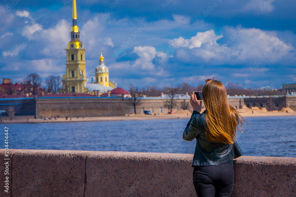 Outdoor view of blonde russian woman taking a picture of a gorgeous Peter and Paul Fortress in the horizont in St. Petersburg