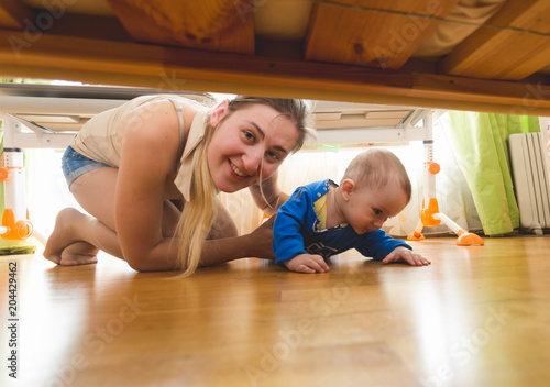 Beautiful young mother with toddler boy lying on floor and looking under the bed