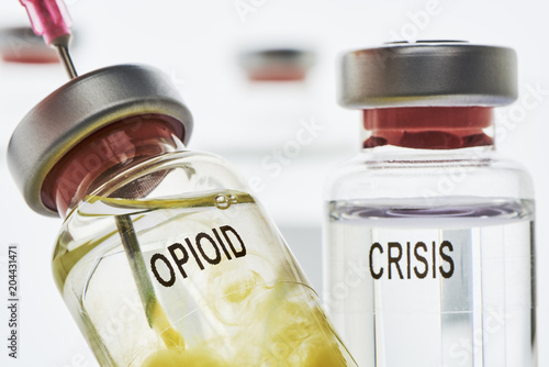 Opioid Krise - In eine Opioid Ampulle die klare flüssigkeit enthält, wird mit einer Nadel eine gelbeflüssigkeit injeziert. Im hintergrund ist ein Ampulle mit der Aufschrift Krise photo