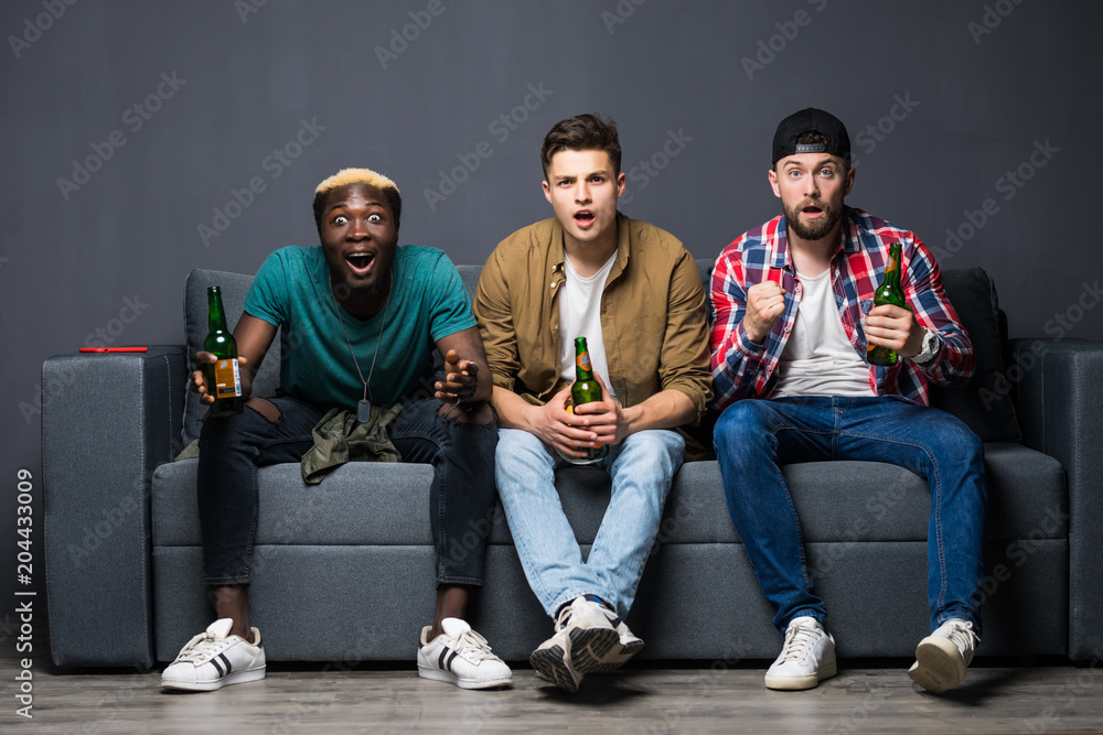 Fototapeta premium Group of exited men sitting together on couch, watching television holding beers, screaming