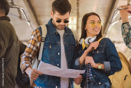 young stylish female traveler in sunglasses with headphones and boyfriend looking at map in metro train