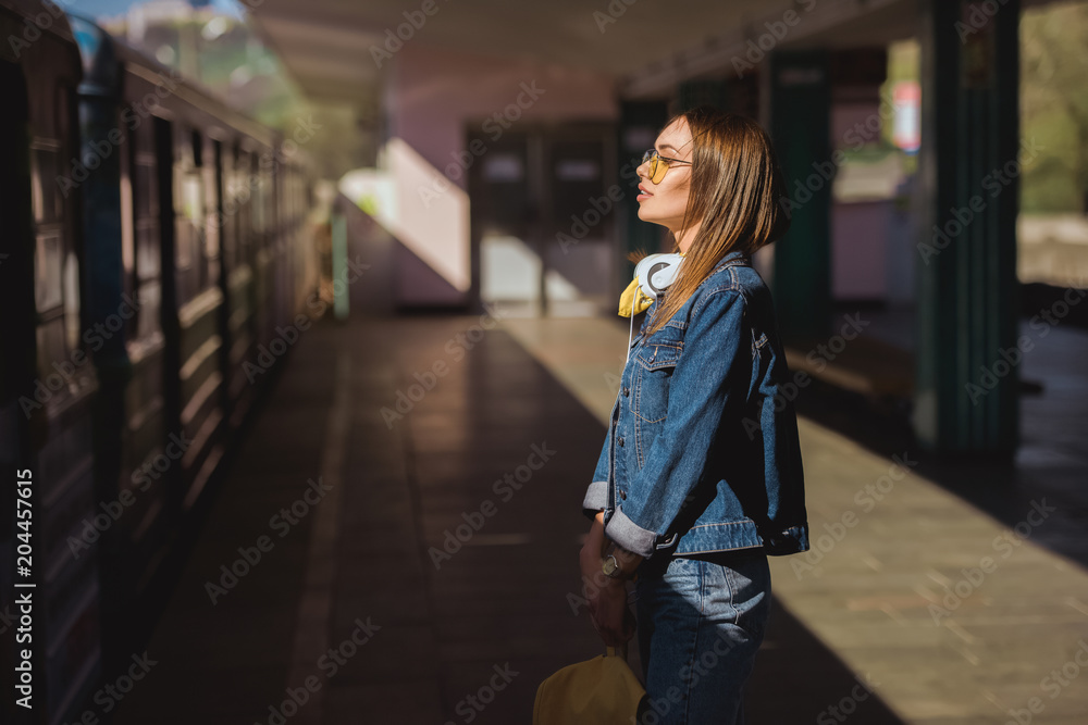 side view of stylish woman in sunglasses with headphones standing at outdoor subway station